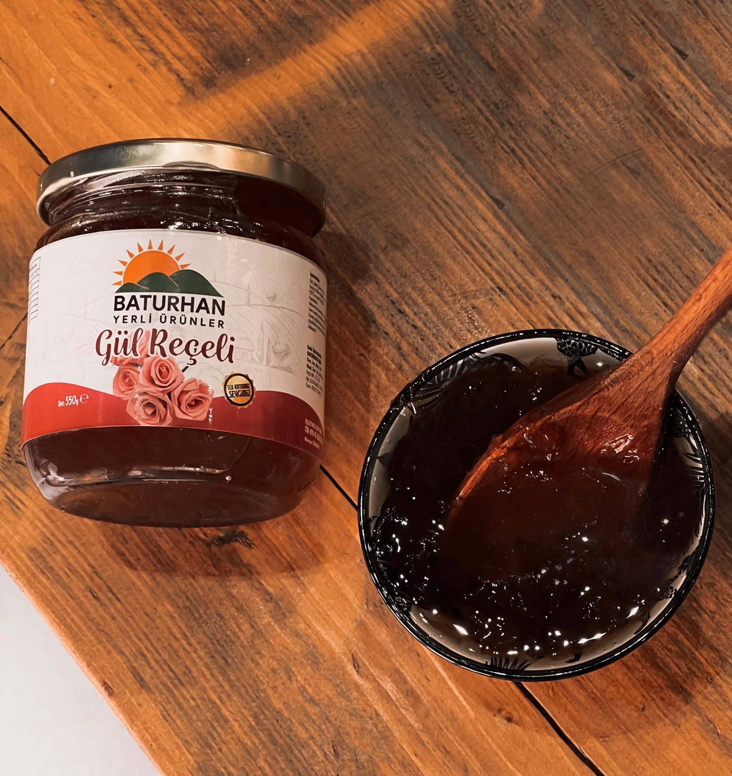 Jampot met Rozenjam (Gul Receli) van Mijn winkel, naast een kom met glanzende rozenjam en een houten lepel, op een rustieke houten tafel.