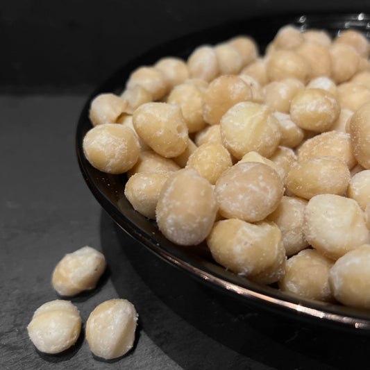 Een zwart bord gevuld met natuurlijke macadamia noten van Mijn winkel. Deze romige en knapperige noten zijn perfect voor een gezonde snack.