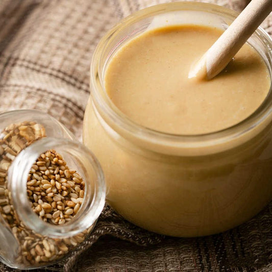 Een pot met tahini dubbel geroosterd van Mijn winkel, gevuld met romige, lichte pasta van geroosterde sesamzaad, afgebeeld met sesamzaadjes op achtergrond.